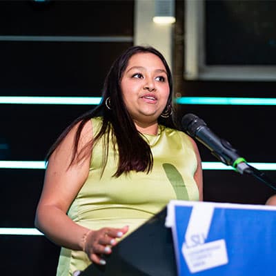 Tanishka is wearing a light green dress and is standing in front of a microphone delivering a speech on stage.